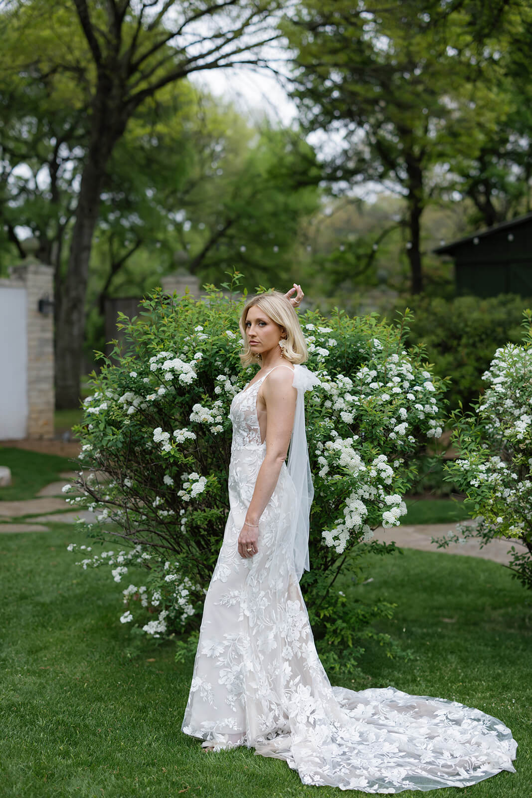 A bride poses in her floral lace wedding gown and long veil beside a large bush covered in white flowers.