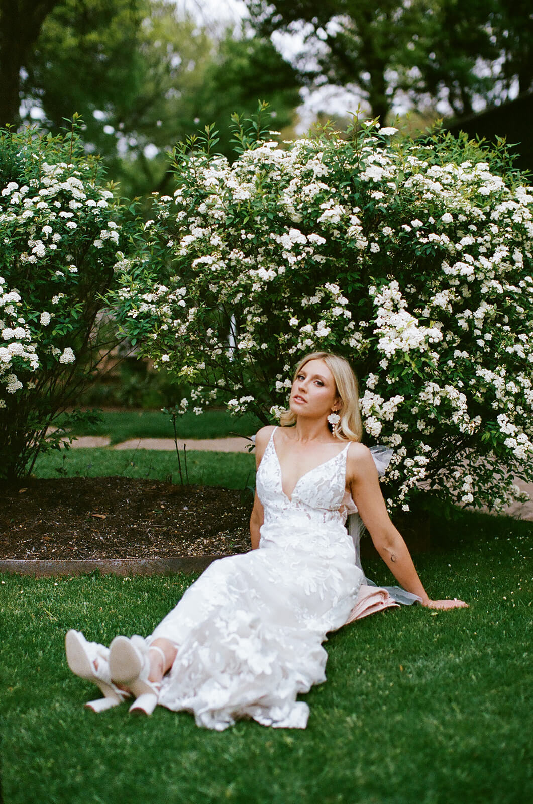 A bride sits on the grass in front of a flowering white rose bush, wearing a floral lace wedding gown and white sneakers.