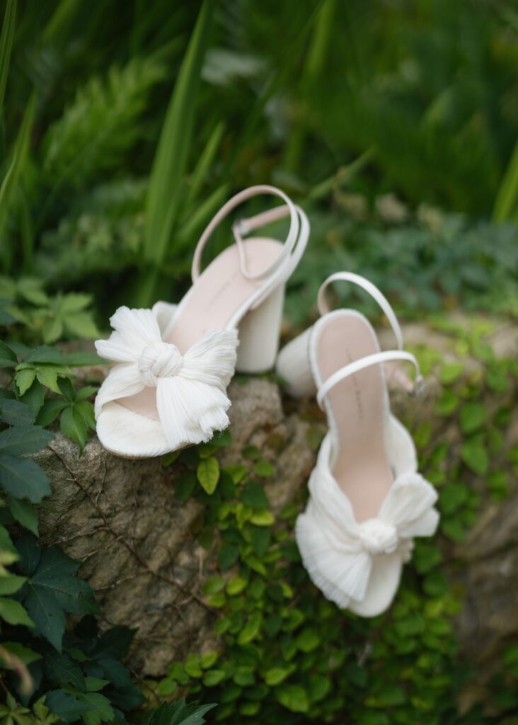 White bridal heels with oversized bows displayed on a moss-covered stone surrounded by garden greenery.