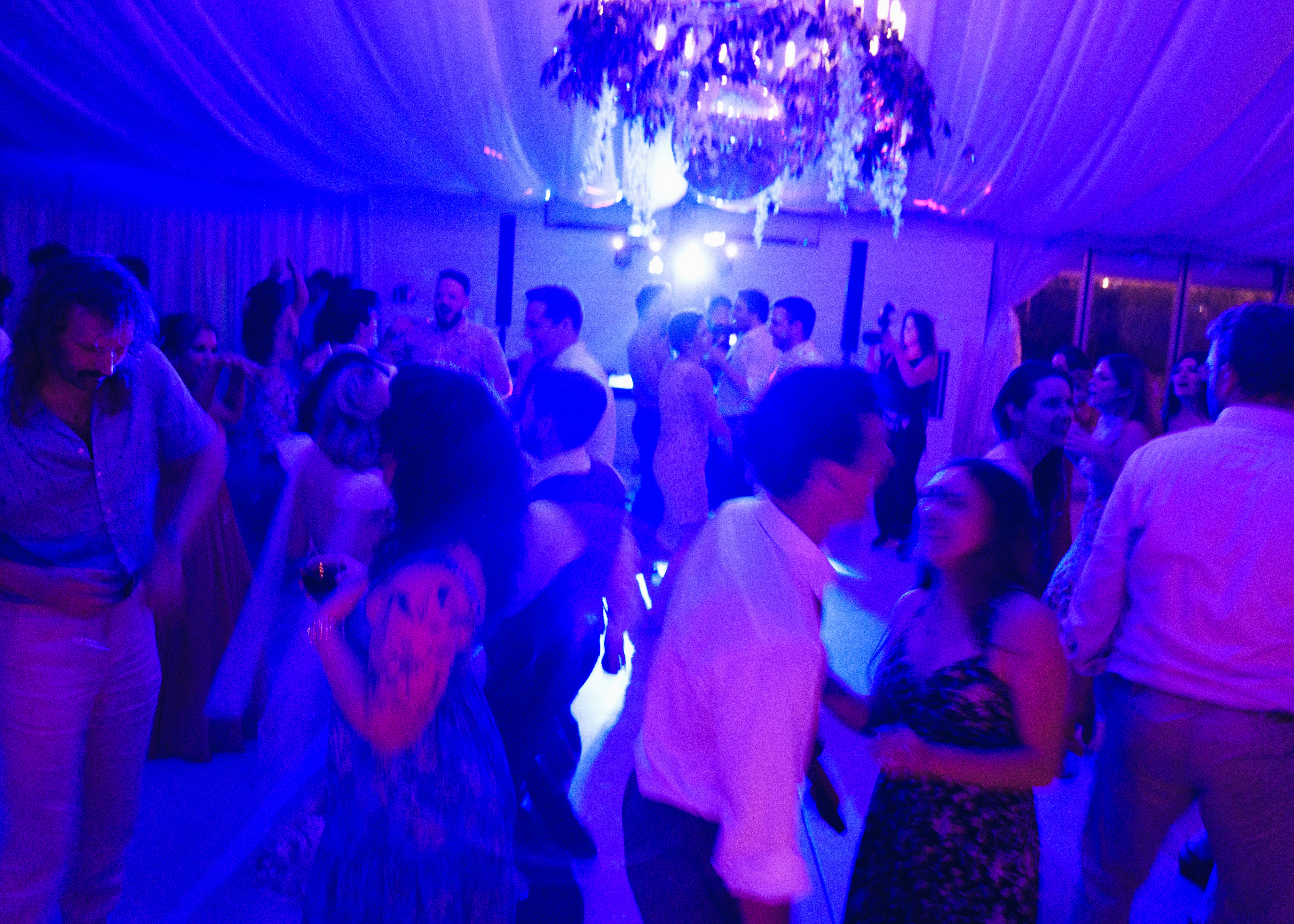 Wedding guests dance beneath colorful light trails and warm reception lighting in an artistic long-exposure photograph.