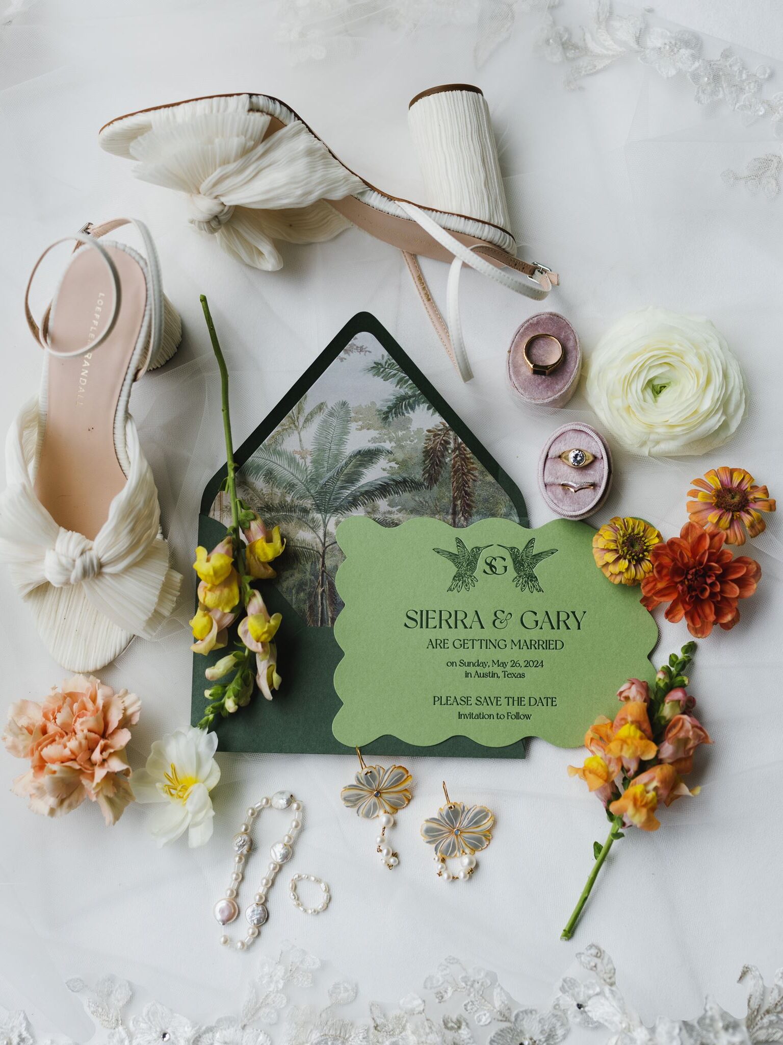 Editorial wedding detail flat lay featuring Sierra and Gary’s green hummingbird save the date, ivory Loeffler Randall bridal heels, velvet ring boxes, pearl jewelry, floral earrings, and colorful garden-inspired blooms arranged on a lace wedding veil at Hummingbird House in Austin, Texas.